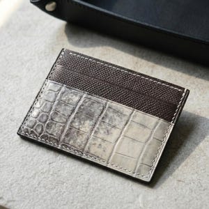 Könnte beinhalten: Ein rechteckiger Kartenhalter mit zweifarbigem Design. Der obere Bereich besteht aus dunkelbraunem, strukturiertem Leder, während der untere Bereich ein hellgraues und weißes Krokodil-Prägemuster mit sichtbaren weißen Nähten aufweist.