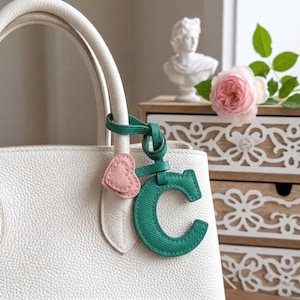 Puede incluir: Un bolso de mano de cuero blanco con una letra "C" de tela verde y un charm en forma de corazón rosa adjunto al asa. El bolso se encuentra sobre un fondo suave con un busto y una rosa rosa.