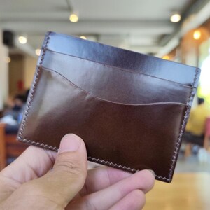 May include: A dark brown leather card holder featuring a smooth, polished finish and visible stitching along the edges. The rectangular accessory includes a curved pocket design on the front, providing a sleek and functional storage solution for cards.