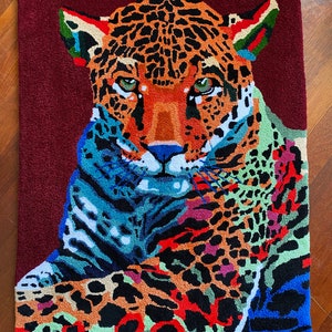 Puede incluir: Una alfombra colorida con un primer plano de la cara de un jaguar. El jaguar tiene un patrón de manchas negras y naranjas con acentos azules, verdes y rojos. La alfombra tiene un fondo burdeos.