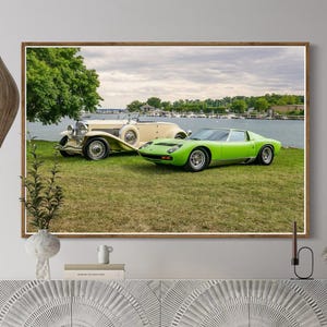 May include: A framed print featuring two classic cars, a vintage cream-colored car and a bright green sports car, parked on a grassy area near a body of water. The print is displayed above a white cabinet and a beige sofa with decorative pillows.