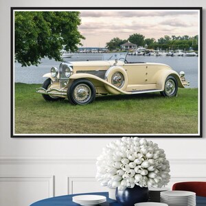 May include: A dining room featuring a framed picture of a vintage cream-colored convertible car. The room has a blue round table with a gold base, red chairs, and a black and white patterned rug. White walls and a large window complete the scene.