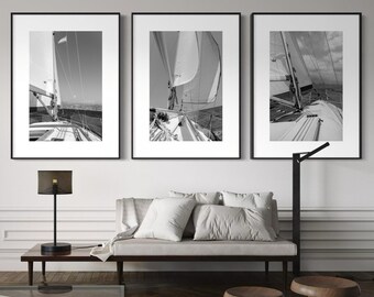 May include: Three framed black and white photographs of sailboats hanging above a gray sofa. The images capture the sails and decks of the boats. The room has a modern design with a glass coffee table, a black lamp, and wooden stools.