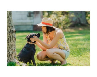 May include: A woman wearing a straw hat with an orange band and a yellow tank top with a floral pattern, petting a black dog wearing a brown collar. The scene takes place outdoors on a sunny day with green grass.