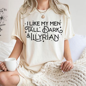 Puede incluir: Camiseta color crema con el texto negro "I like my men tall, dark & Illyrian". La camiseta tiene un corte holgado y mangas cortas. Una taza blanca se sostiene en la mano de la persona.