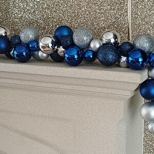 May include: A decorative garland of Christmas ornaments in shades of blue, silver, and glittery silver. The ornaments are arranged on a white mantelpiece against a sparkly gold background. The garland includes various sizes and finishes.