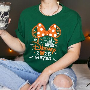 May include: Green t-shirt featuring a Halloween-themed Disney design. The design includes an orange polka-dot bow, a castle, and the text "Disney 2025 Sister". The shirt is made of a soft material.