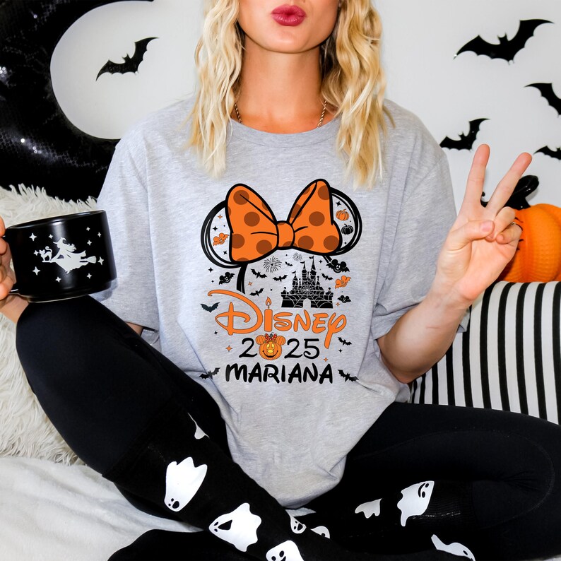 May include: A light gray t-shirt with a Halloween-themed Disney design. The design includes a large orange bow with polka dots, Mickey Mouse ears, a castle, and the text "Disney 2025 MARIANA". The person is also holding a black mug.