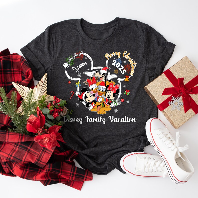 May include: Dark gray t-shirt featuring a Disney family vacation design with Mickey, Minnie, Donald, Daisy, and Goofy in Santa hats. The shirt has the text "Merry Christmas 2025" and "Disney Family Vacation". A pair of white sneakers and a wrapped gift are also in the image.