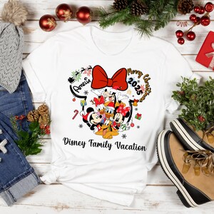 May include: White t-shirt featuring a Disney family graphic with Mickey, Minnie, Donald, Daisy, and Goofy. The design includes a red bow, the words "Merry Christmas 2025", and "Disney Family Vacation".