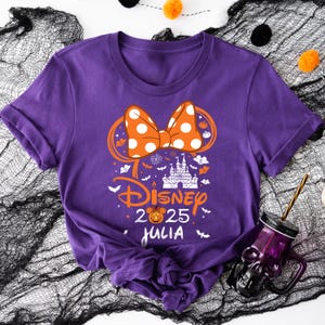 May include: Purple t-shirt featuring a Disney-themed design with a polka-dot bow, castle, and the text "Disney 2025 Julia". The design includes pumpkins and bats, suggesting a Halloween theme.