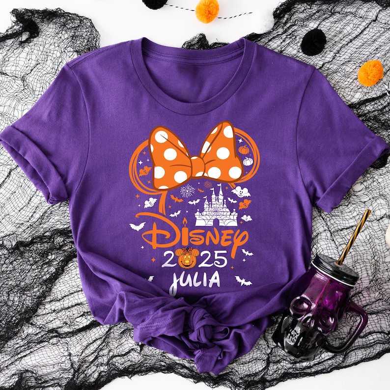 May include: Purple t-shirt featuring a Disney-themed design with a polka-dot bow, castle, and the text "Disney 2025 Julia". The design includes pumpkins and bats, suggesting a Halloween theme.