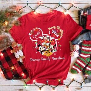 May include: A red t-shirt featuring a Disney family graphic with Mickey, Minnie, Donald, Daisy, Goofy, and Pluto. The shirt has the text "Merry Christmas 2025" and "Disney Family Vacation".