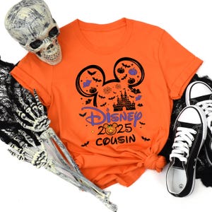 May include: Orange t-shirt with a Disney Halloween design. The design includes Mickey Mouse ears, a castle, pumpkins, bats, and the text "Disney 2025 Cousin". Black and white sneakers are visible on the right.