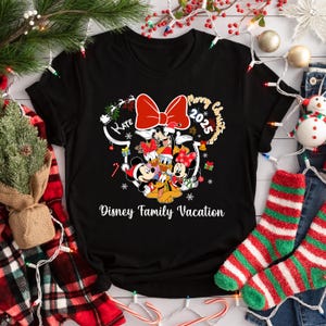 May include: Black t-shirt featuring a festive Disney design. The design includes Mickey and Minnie Mouse, Donald and Daisy Duck, and Goofy, with a large red bow and the text "Merry Christmas 2025" and "Disney Family Vacation".