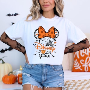 May include: White t-shirt featuring a Halloween-themed Disney design. The design includes an orange polka-dot bow, Mickey Mouse ears, a castle silhouette, and the text "Disney 2025 Lydia" in orange and black. Bats and pumpkins are also included.