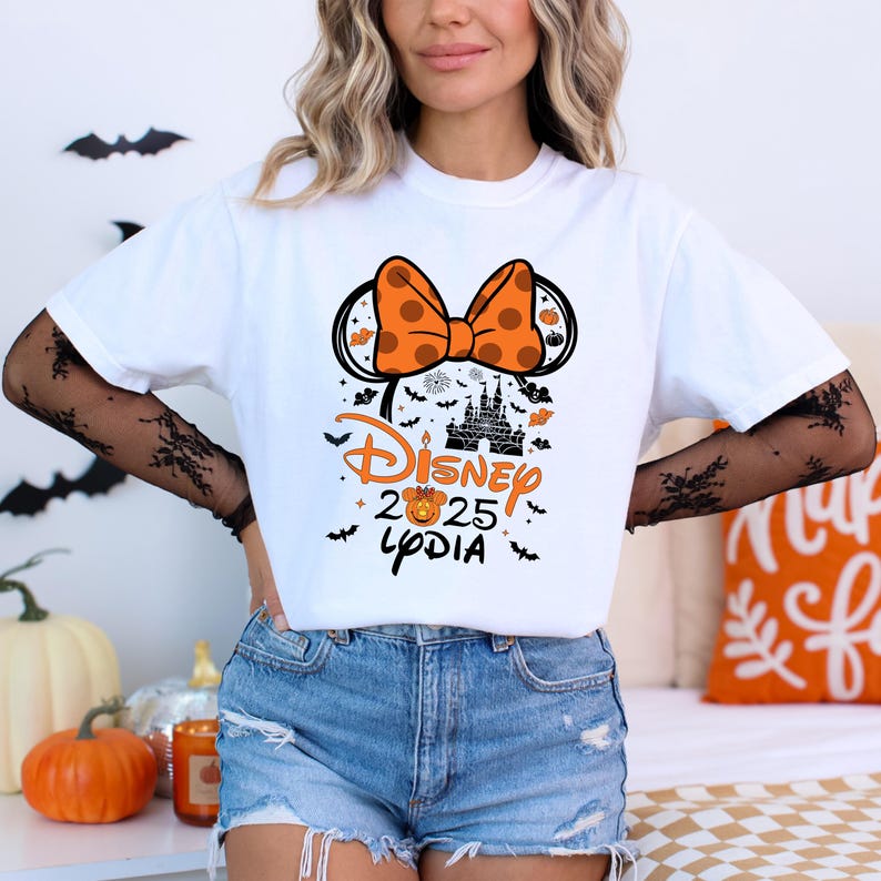 May include: White t-shirt featuring a Halloween-themed Disney design. The design includes an orange polka-dot bow, Mickey Mouse ears, a castle silhouette, and the text "Disney 2025 Lydia" in orange and black. Bats and pumpkins are also included.