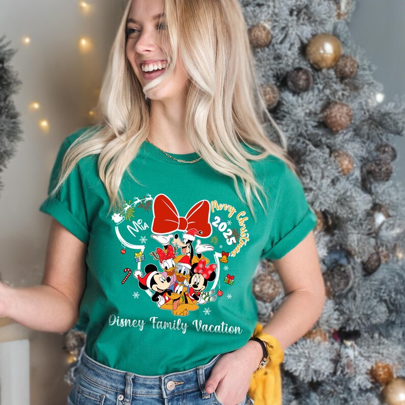 May include: Green t-shirt with a festive design featuring Disney characters, a red bow, and the text "Merry Christmas 2025" and "Disney Family Vacation". The shirt has rolled-up sleeves.
