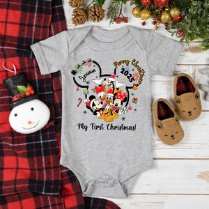 May include: A gray baby onesie featuring Mickey Mouse and friends with the text "Merry Christmas 2025" and "My First Christmas!". The design includes a name and festive graphics. Two brown baby shoes are next to the onesie.