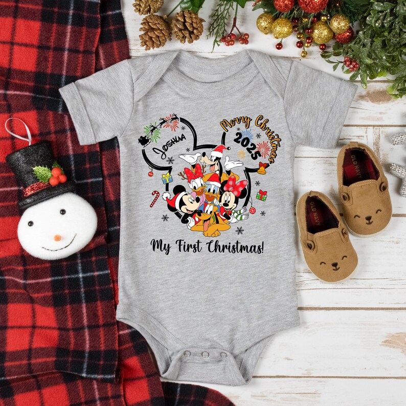May include: A gray baby onesie featuring Mickey Mouse and friends with the text "Merry Christmas 2025" and "My First Christmas!". The design includes a name and festive graphics. Two brown baby shoes are next to the onesie.