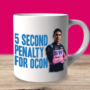 Puede incluir: Taza de cerámica blanca con un piloto de carreras en azul y rosa. El texto "5 SECOND PENALTY FOR OCON" está impreso en azul en la taza.