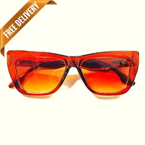 Puede incluir: Gafas de sol rojas con forma de ojo de gato y lentes degradadas naranjas. Las gafas de sol tienen una montura gruesa y transparente y una forma clásica. La imagen incluye una pancarta dorada con el texto "FREE DELIVERY".