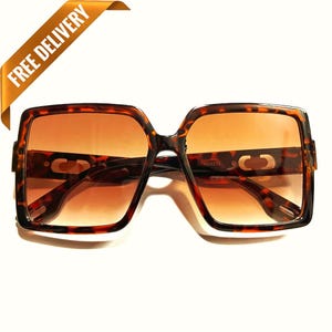 Puede incluir: Gafas de sol cuadradas con estampado de carey. Las lentes son de color marrón degradado. Las gafas de sol tienen una montura gruesa y un diseño clásico. La imagen también incluye una pancarta dorada que dice "FREE DELIVERY".
