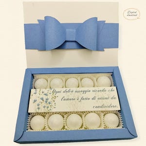 May include: A light blue paper gift box with a large bow on the lid, containing ten white spherical sweets in paper liners. A rectangular card sits in the centre with blue floral illustrations and the Italian text: Ogni dolce assaggio ricorda che l'amore è fatto di attimi da condividere.