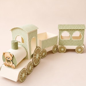 Puede incluir: Un tren de papel en color verde salvia y crema, que incluye una locomotora con una ilustración de un osito de peluche, un vagón de carga y un vagón de pasajeros. El tren presenta patrones de cuadros vichy y lunares, con ventanas y ruedas recortadas.