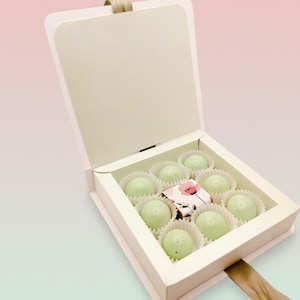 May include: A square, light pink gift box with an open lid containing eight round, pale green candies in white paper liners. A small, rectangular decorative paper sleeve with a pink floral pattern sits in the center among the candies.