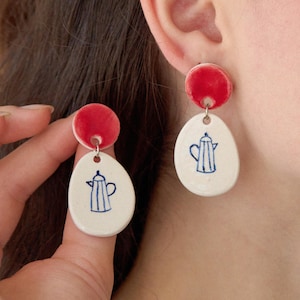 May include: A pair of ceramic earrings with a red circle and a white oval with a blue and white teapot design.