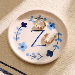 Könnte beinhalten: Eine weiße Keramik-Schmuckschale mit einem blauen Blumendesign und dem Buchstaben "Z" in der Mitte. Zwei goldene Creolen liegen auf der Schale.