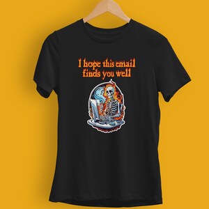 Peut inclure: T-shirt noir avec un motif d'un squelette devant un ordinateur, entouré de flammes. Le texte orange au-dessus dit "I hope this email finds you well". Le t-shirt est suspendu à un cintre en bois.