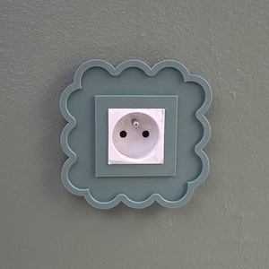 Peut inclure: Une prise murale décorative de couleur bleu-vert. Le cache a une forme festonnée en forme de nuage entourant un cadre carré. La prise elle-même est blanche avec deux trous et une broche de mise à la terre.