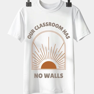 Może przedstawiać: Biała koszulka z brązowym napisem "OUR CLASSROOM HAS NO WALLS". Projekt przedstawia brązowy łuk z grafiką promieni słonecznych. Koszulka wisi na czarnym klipsie.