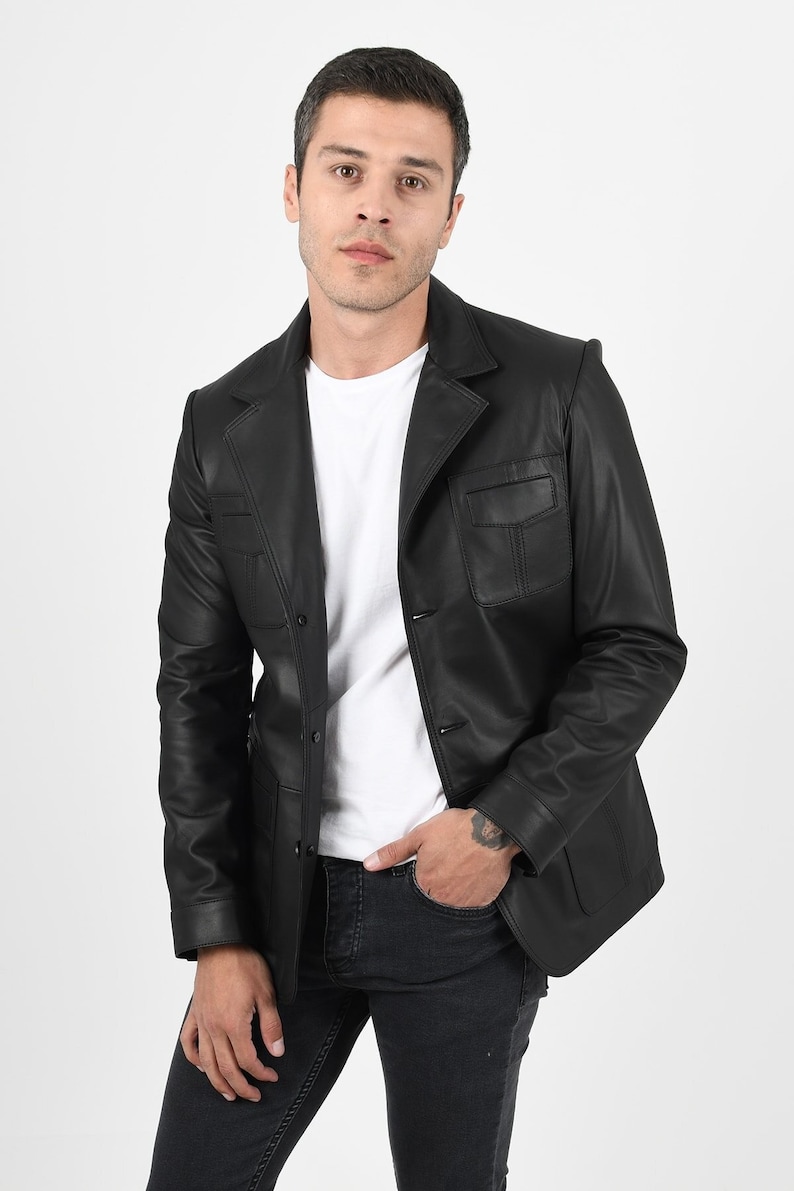 May include: A black leather blazer with a notched lapel, two front pockets, and a two-button closure. The blazer is worn over a white t-shirt and paired with black jeans. The model is standing in front of a white background.