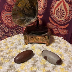 May include: A miniature antique gramophone with a copper finish and a large horn. Two oval-shaped stones, one brown and one gray with a brown spot, are in front of the gramophone. The background is a patterned purple fabric.