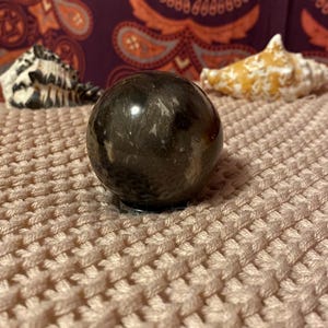 May include: A black obsidian sphere with white and gray swirls. The sphere is sitting on a light pink knitted surface.