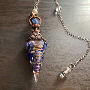 Puede incluir: Un péndulo de cristal de cuarzo transparente con un alambre de cobre envuelto alrededor. El cristal está lleno de piedras azules y blancas. El péndulo está unido a una cadena plateada con un cristal transparente en el extremo.