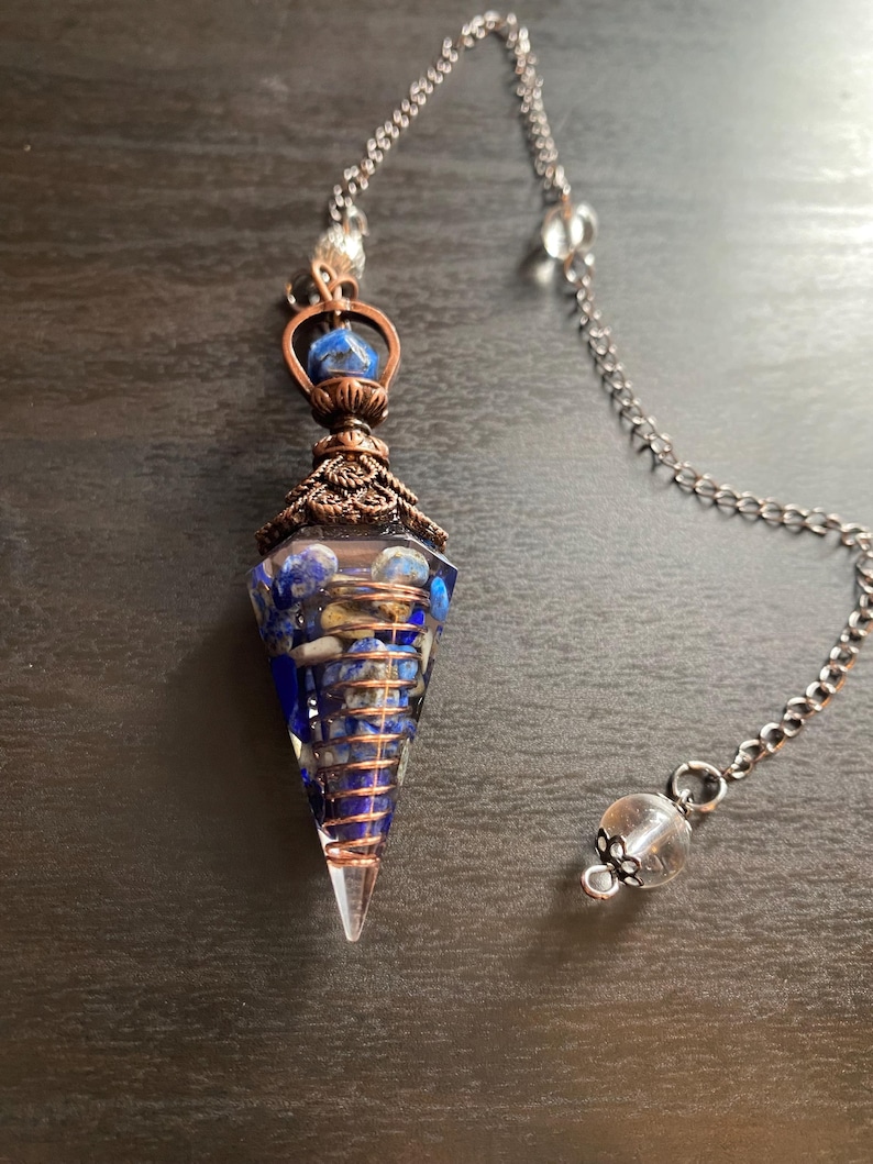 Op de afbeelding: Een helder kwartskristal slinger met een koperen draad eromheen gewikkeld. Het kristal is gevuld met blauwe en witte stenen. De slinger is bevestigd aan een zilveren ketting met een helder kristal aan het einde.
