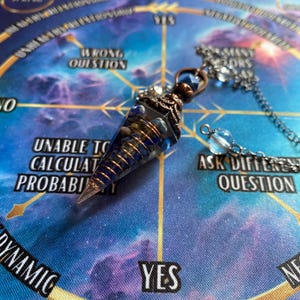 Op de afbeelding: Een blauwe en koperen slinger met een kristallen punt, hangend aan een ketting, boven een cirkelvormig diagram met de woorden "JA", "NEE", "VERKEERDE VRAAG", "ONVERMOGEN OM DE WAARSCHIJNLIJKHEID TE BEREKENEN", "ANDERE VRAAG STELLEN", "DYNAMISCH", en "TEGEN DE KLOK IN DRAAIEN".