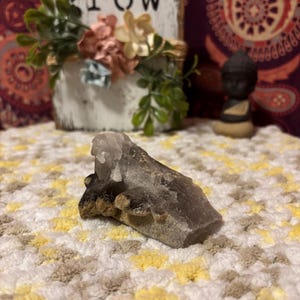 May include: A smoky quartz crystal with a dark gray and clear color variation. The crystal is resting on a crocheted surface.