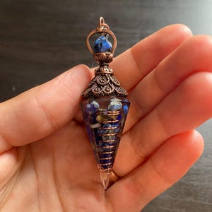Op de afbeelding: Een met koperdraad gewikkelde kristallen punt hanger met een blauwe en gouden steen aan de bovenkant. De hanger is gevuld met blauwe en gouden stenen en hangt aan een koperen ketting.