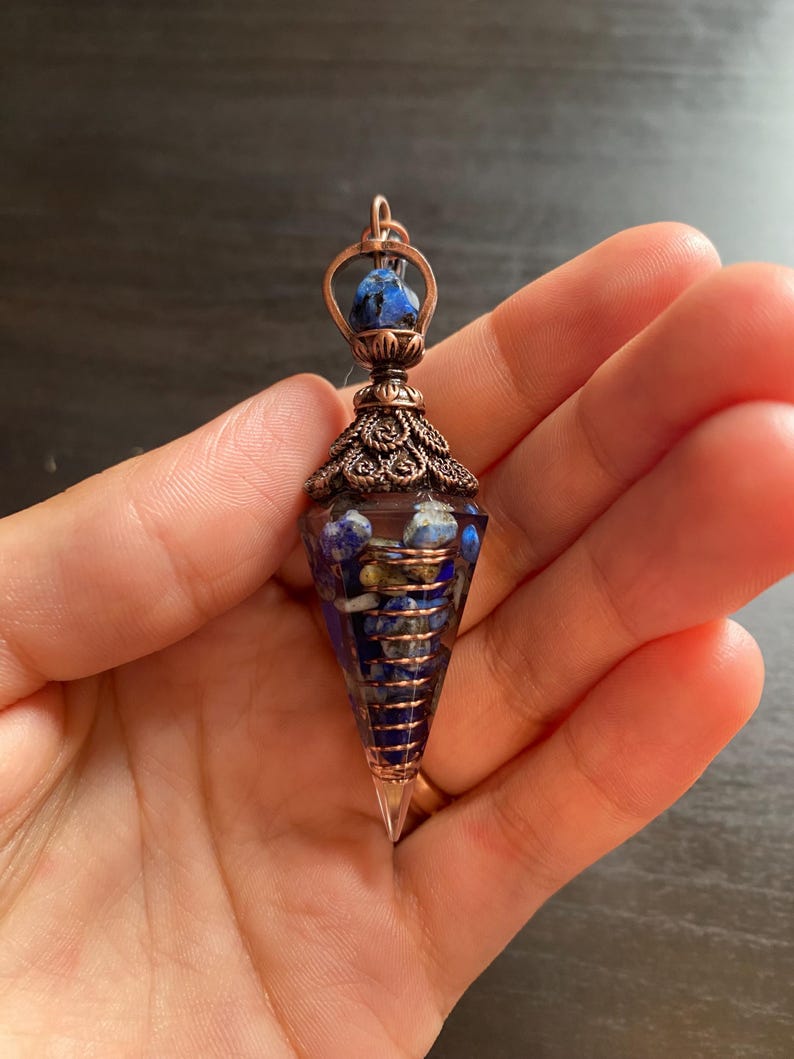 Op de afbeelding: Een met koperdraad gewikkelde kristallen punt hanger met een blauwe en gouden steen aan de bovenkant. De hanger is gevuld met blauwe en gouden stenen en hangt aan een koperen ketting.