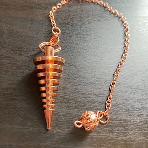 Puede incluir: Un péndulo de color cobre con un diseño en espiral y una cadena. El péndulo está unido a una pequeña bola redonda decorativa.