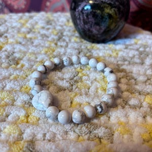 May include: White Howlite beaded bracelet with a heart-shaped bead. The bracelet is on a white and yellow textured surface.