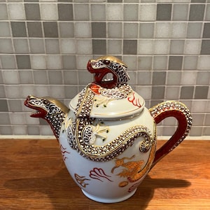 Peut inclure: Théière en porcelaine blanche avec un motif de dragon. La théière a un dragon rouge et or sur le couvercle et le corps. La théière est décorée d'accents dorés et blancs.