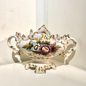 Op de afbeelding: Een witte keramische vaas met twee handvatten en een bloemenontwerp. De vaas heeft een gegolfde rand en een voet met drie poten. De bloemen zijn geschilderd in tinten roze, geel, blauw, groen en bruin.