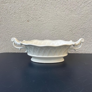 Pode incluir: Vaso de cerâmica branco com duas alças em forma de volutas. O vaso tem uma borda ondulada e um padrão decorativo em relevo.