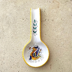 May include: White ceramic spoon rest with a yellow border and handle. The spoon rest features a colorful illustration of a mythical creature and green leaf design. A decorative kitchen accessory.